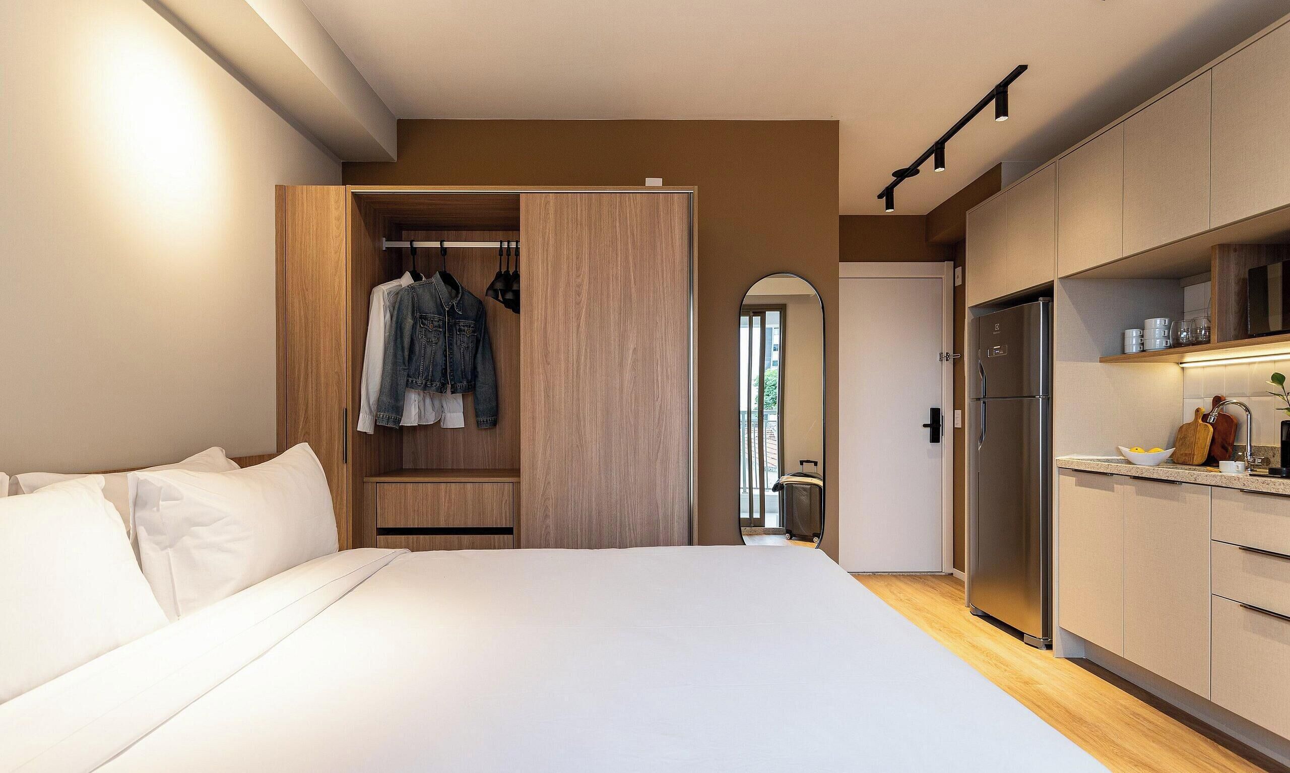 A bedroom with a bed, a wooden wardrobe, and a kitchenette with a refrigerator and a sink.