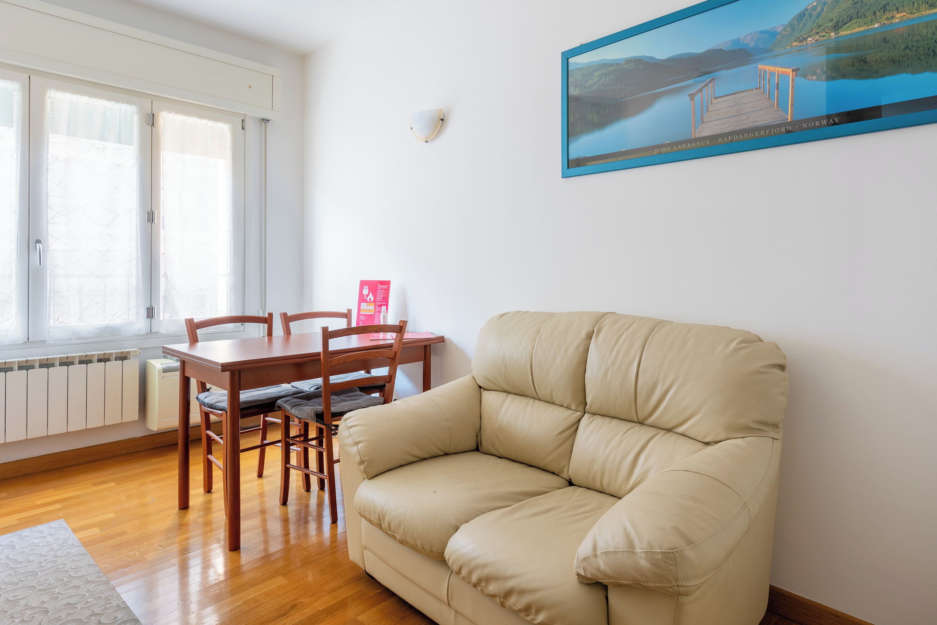 A living room with a beige sofa, wooden dining table, and chairs. A framed picture hangs on the wall.