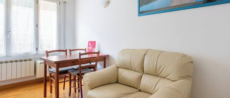 A living room with a beige sofa, wooden dining table, and chairs. A framed picture hangs on the wall.