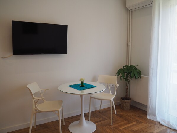 A room with a flat-screen TV mounted on the wall, a round glass table with two white chairs, a small potted plant, and a window with sheer curtains.