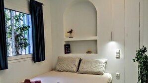 A neatly made bed with rolled towels, a bookshelf with books and a decorative item, a window with curtains, and a potted plant. - Athens White House 🏠  (Athina)