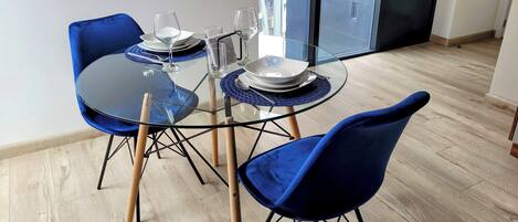 A dining area with a round glass table, blue velvet chairs, and a modern kitchen in the background.