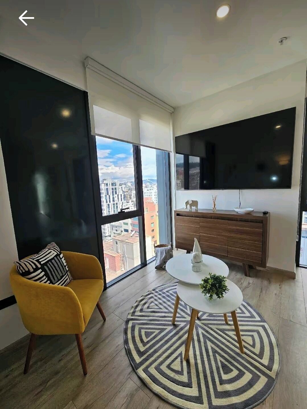 A modern living room with a large window, a yellow armchair, a round coffee table, and a wooden cabinet.