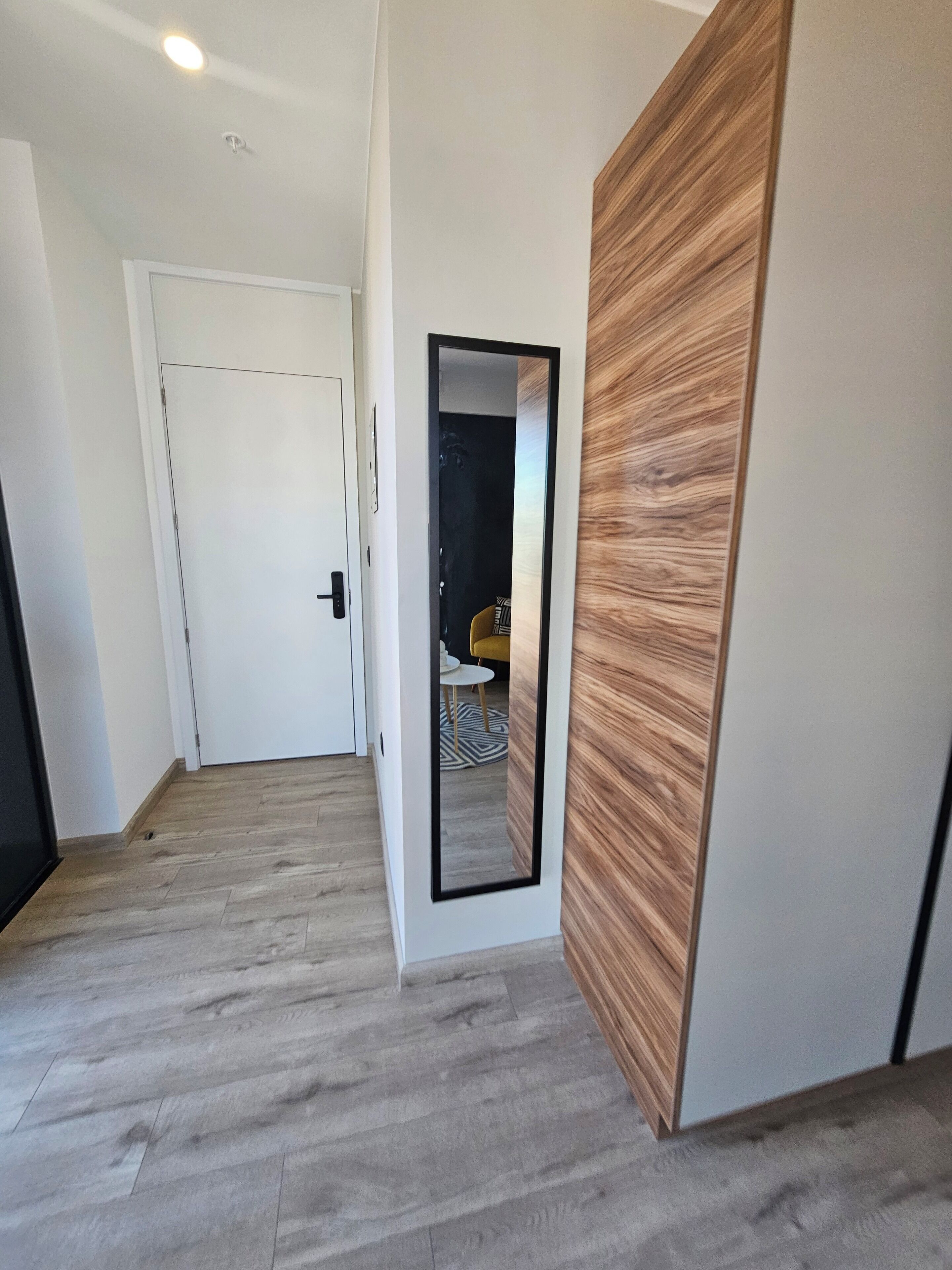 A hallway with a wooden floor, a white door, and a mirror reflecting a room with a yellow chair and a patterned rug.