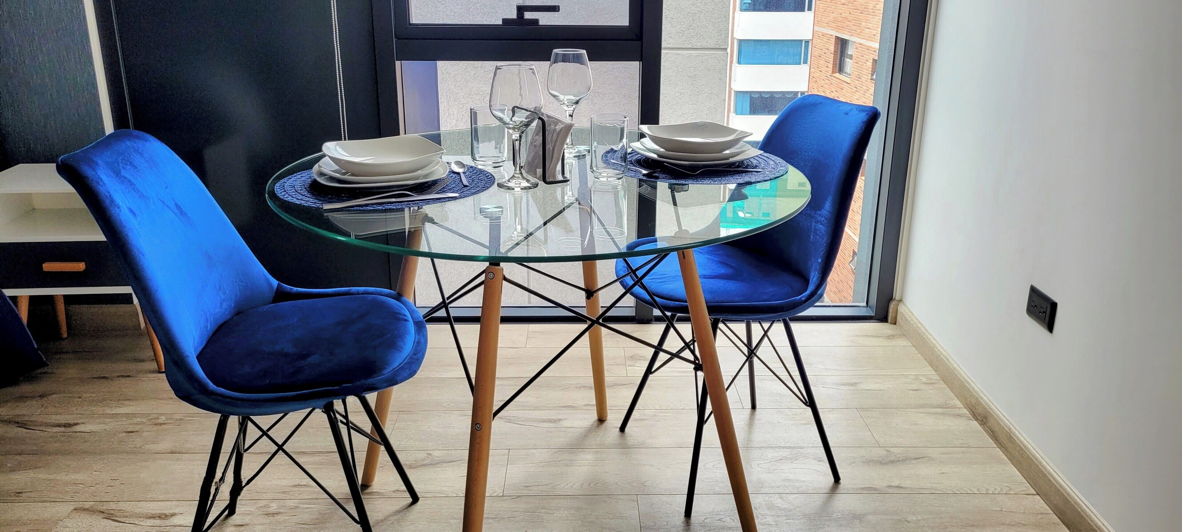 A dining area with a glass table and blue chairs, a small side table with a glass and a plate, and a view of buildings through a window.