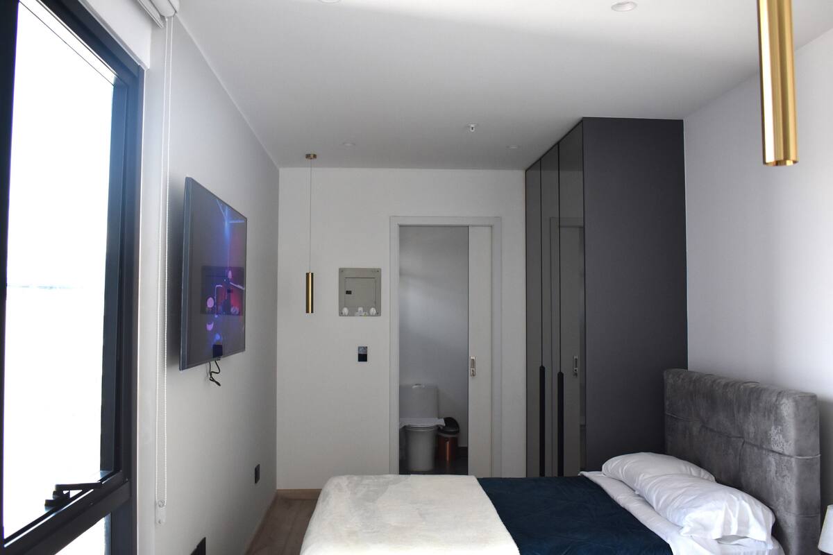 A modern hotel room with a bed, a TV mounted on the wall, a wardrobe, and a window.