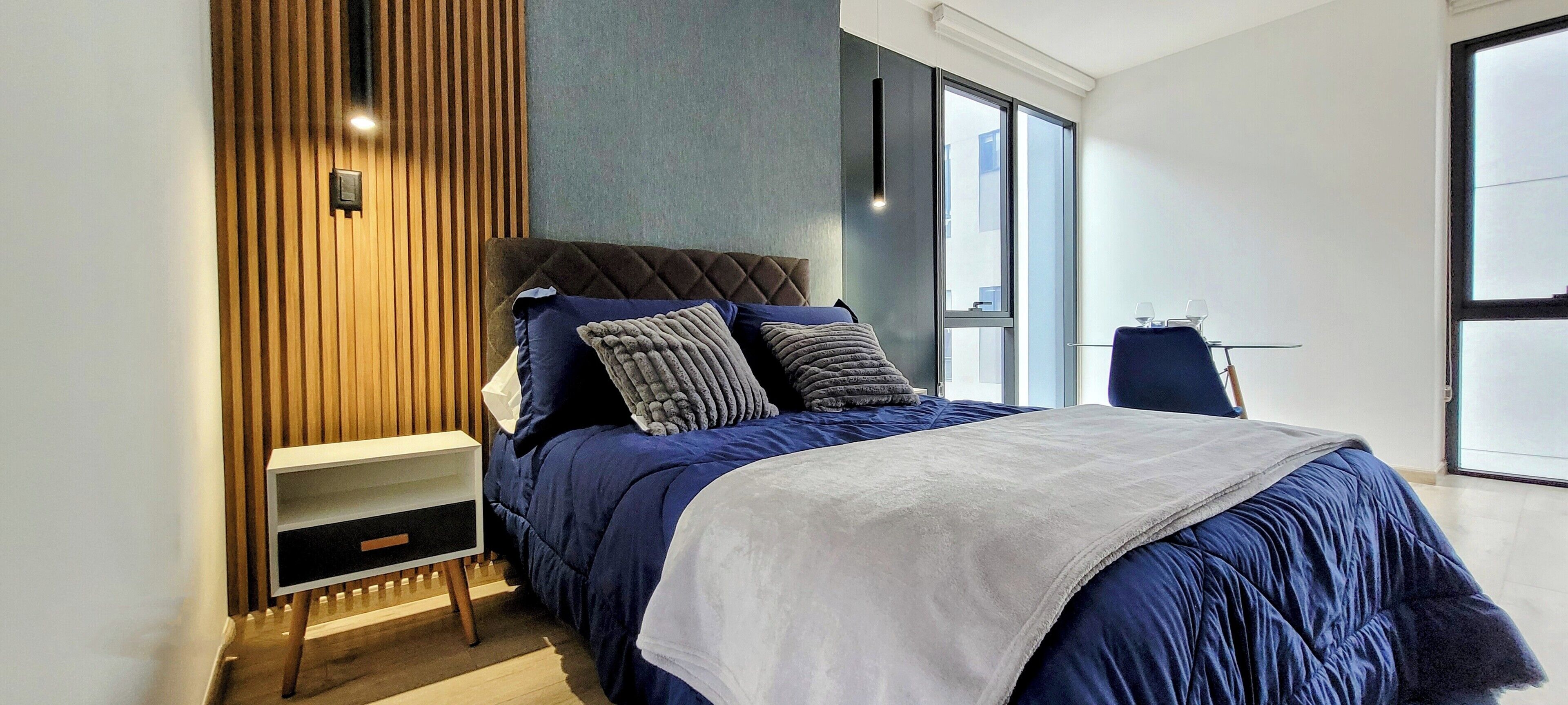 A modern bedroom with a large bed, a nightstand, a wall-mounted light, and a view of the city through a large window.