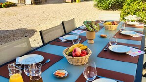 Un espacio de comedor al aire libre con mesas dispuestas para dos, destacando una cesta de fruta, copas de vino y una botella de jugo.