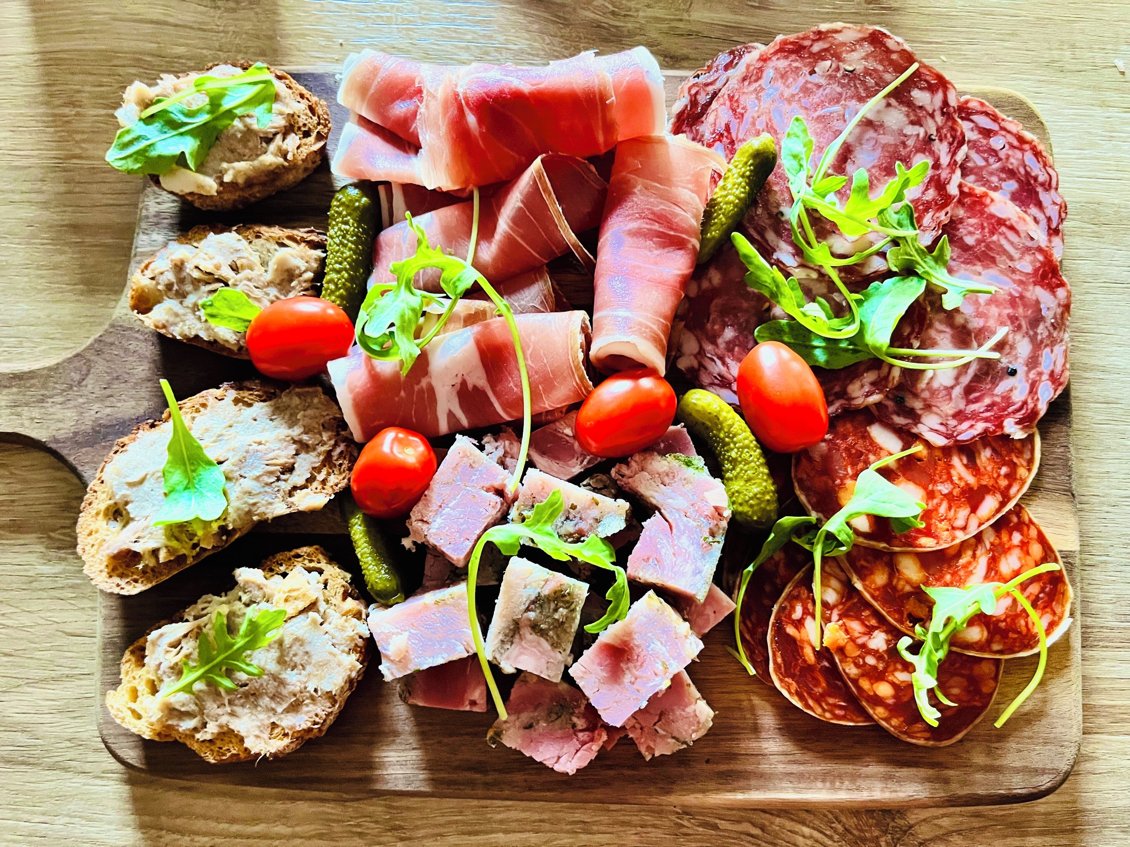 A wooden board with various meats, pickles, tomatoes, and greens.