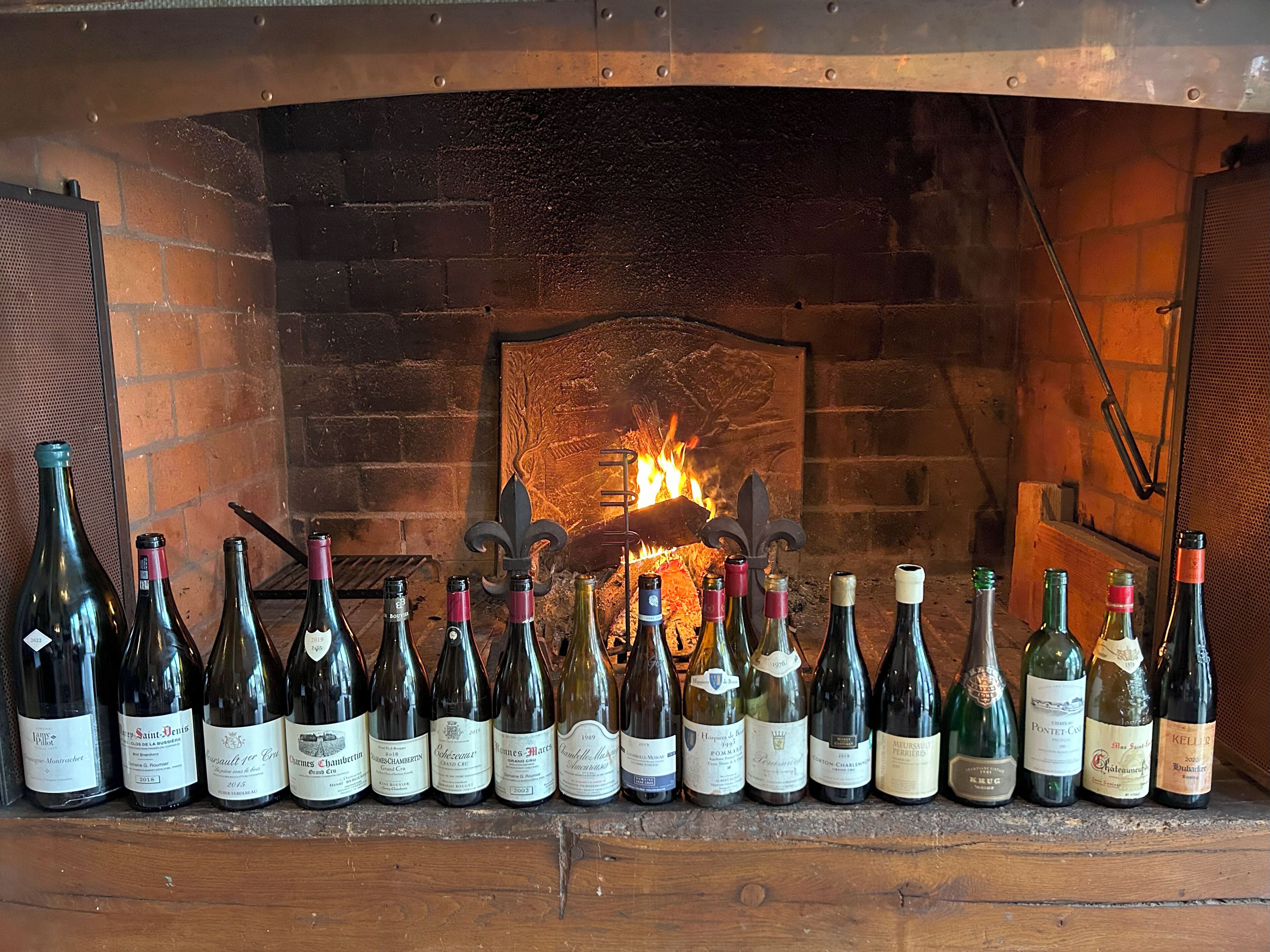A row of wine bottles in front of a brick fireplace with a fire burning.