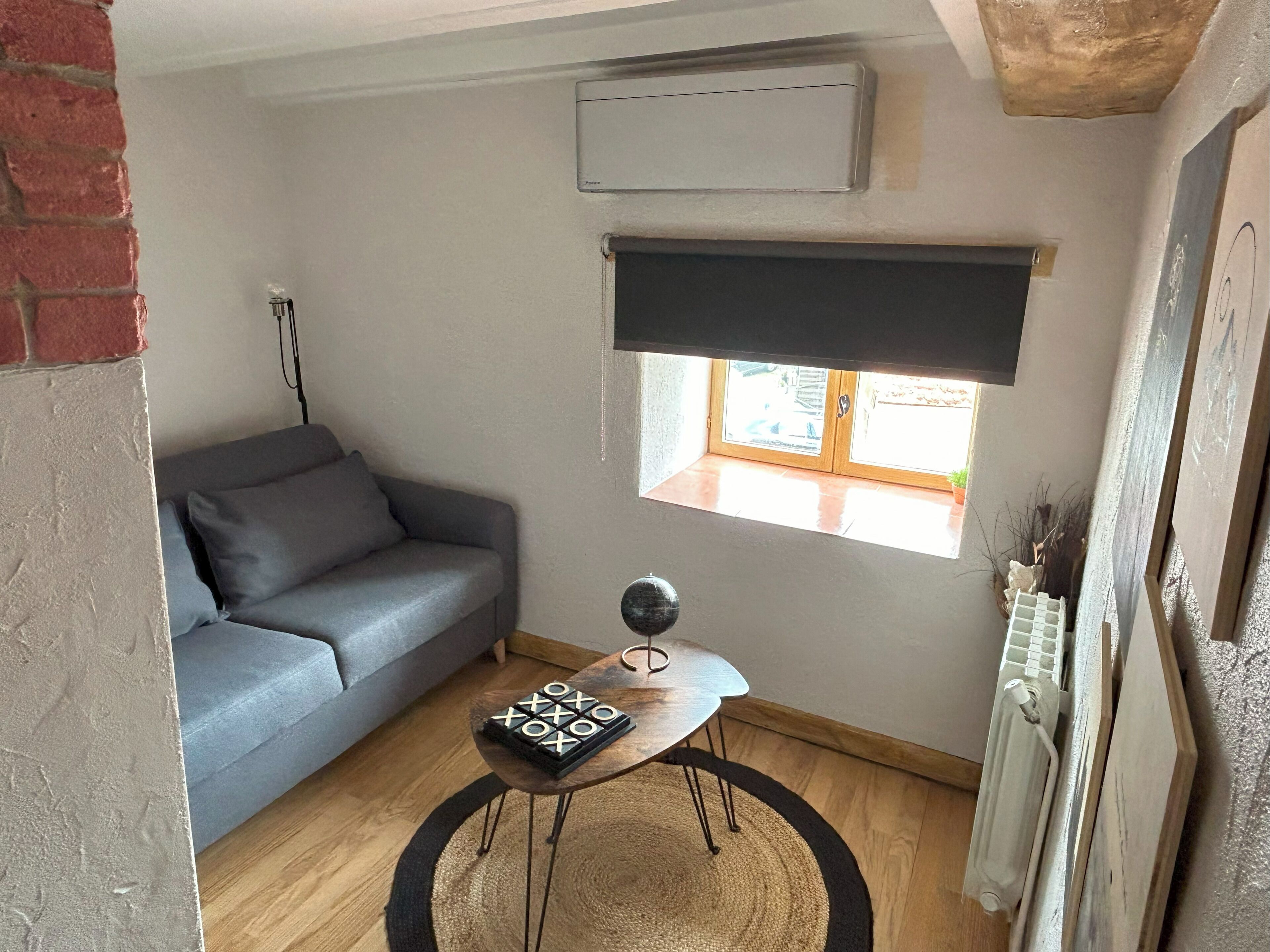 Una habitació petita i acollidora amb un sofà gris, una taula de fusta petita amb un joc de taula, una finestra amb una persiana i un radiador.