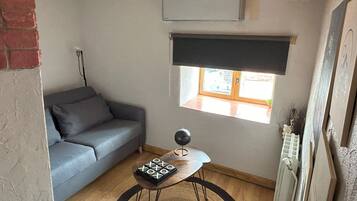 Una habitació petita i acollidora amb un sofà gris, una taula de fusta petita amb un joc de taula, una finestra amb una persiana i un radiador.