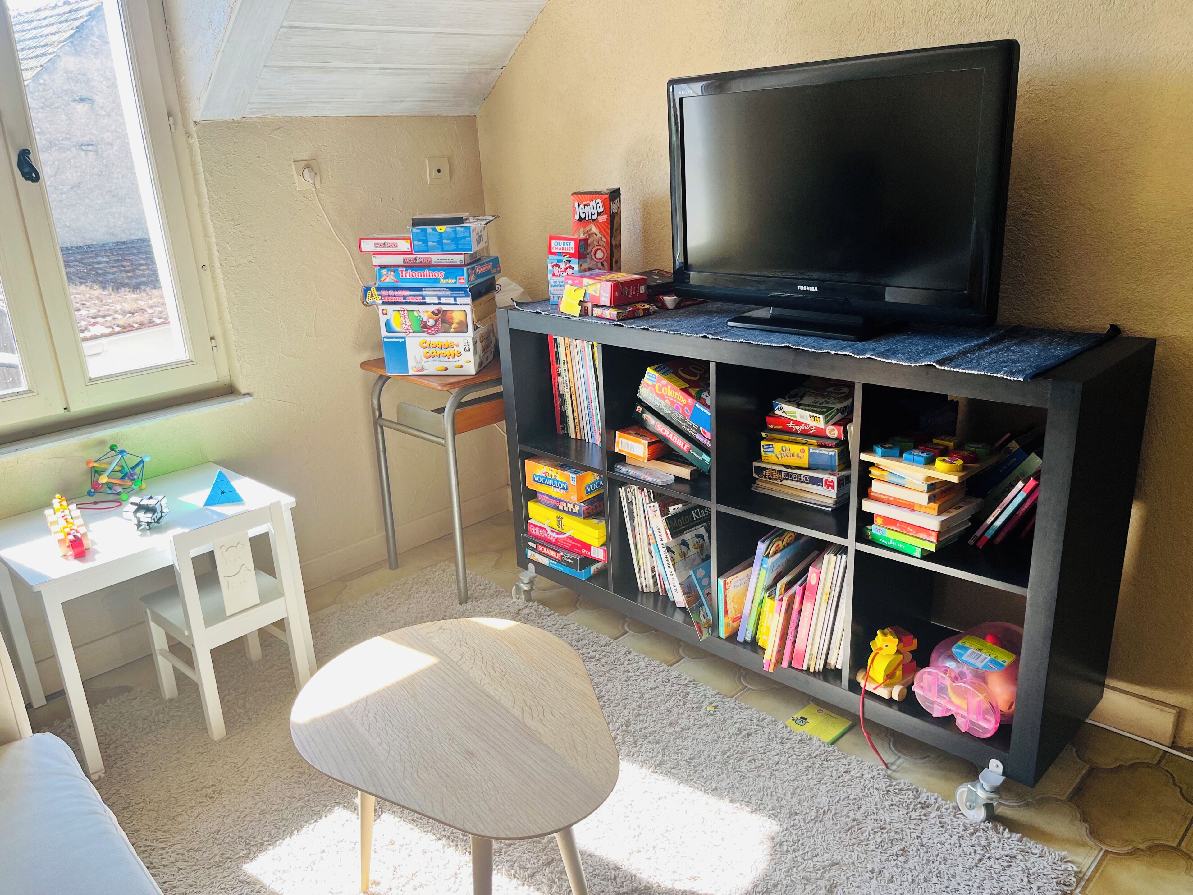 A room with a television on a stand, a bookshelf filled with books and toys, a small white table with a chair, and a window with a view of the outdoors.