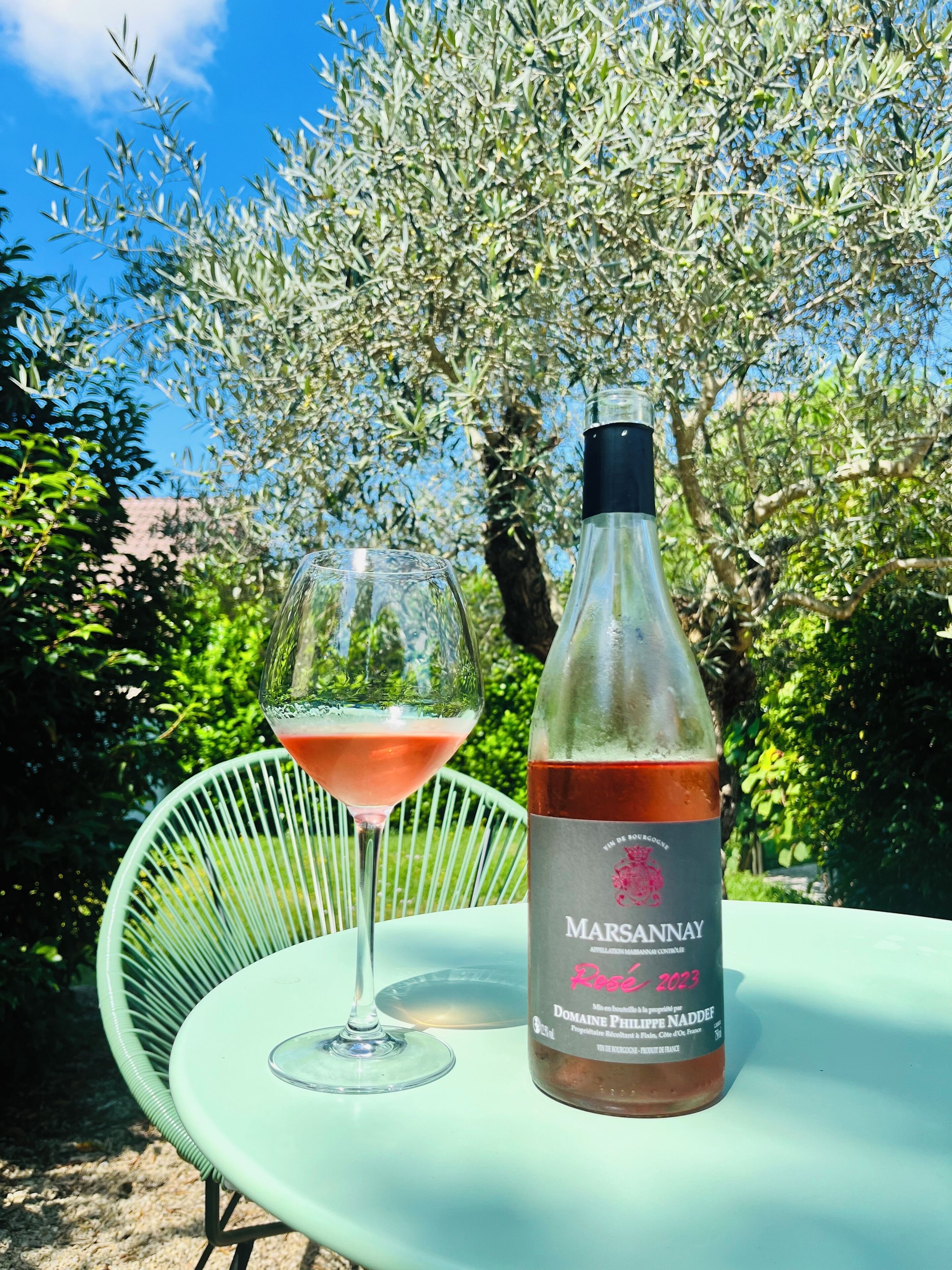 A bottle of Marsannay Rose 2023 wine and a glass of rosé wine on a green table outdoors.