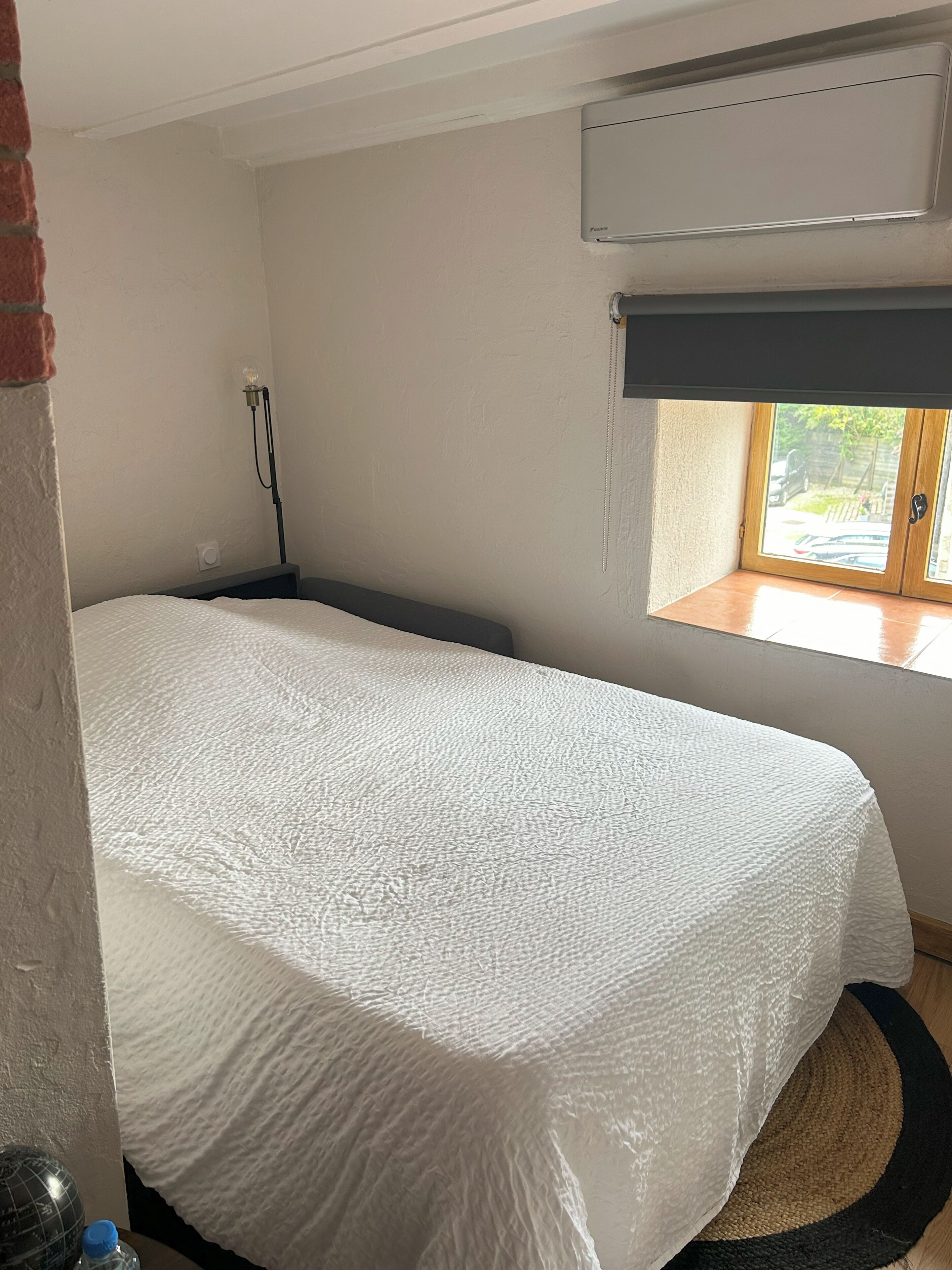 A bedroom with a bed, a window, an air conditioning unit, and a lamp.