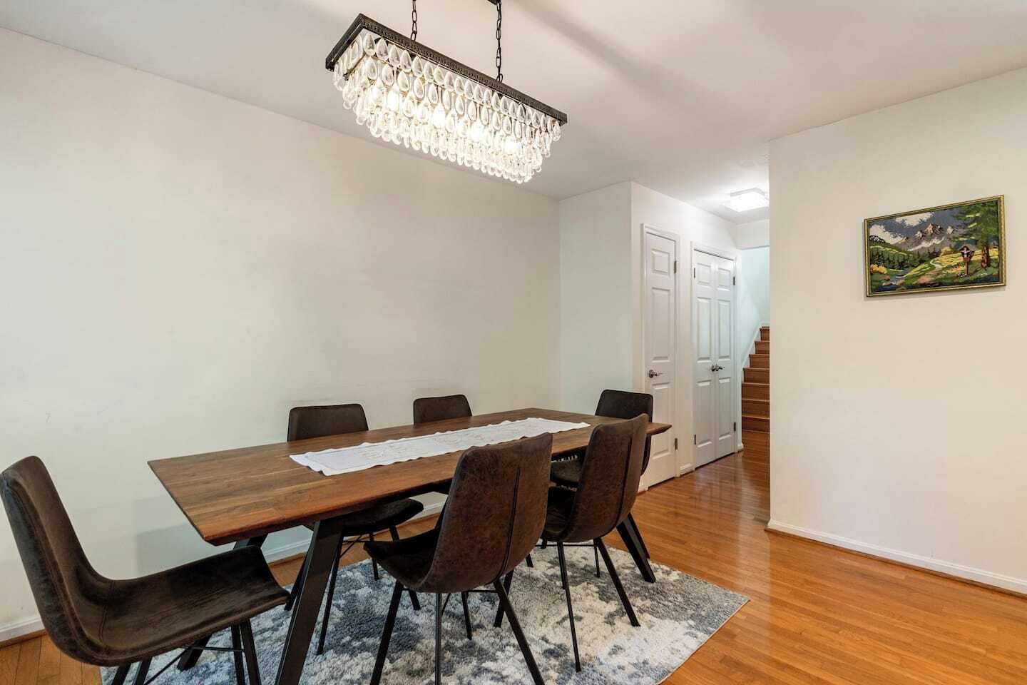 Un coin repas avec une table en bois, six chaises, un lustre, un tapis et un tableau accroché au mur.