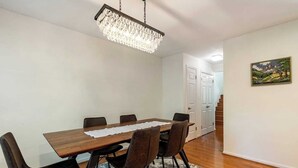 Een eetgedeelte met een houten tafel, zes stoelen, een kroonluchter, een vloerkleed en een schilderij aan de muur.