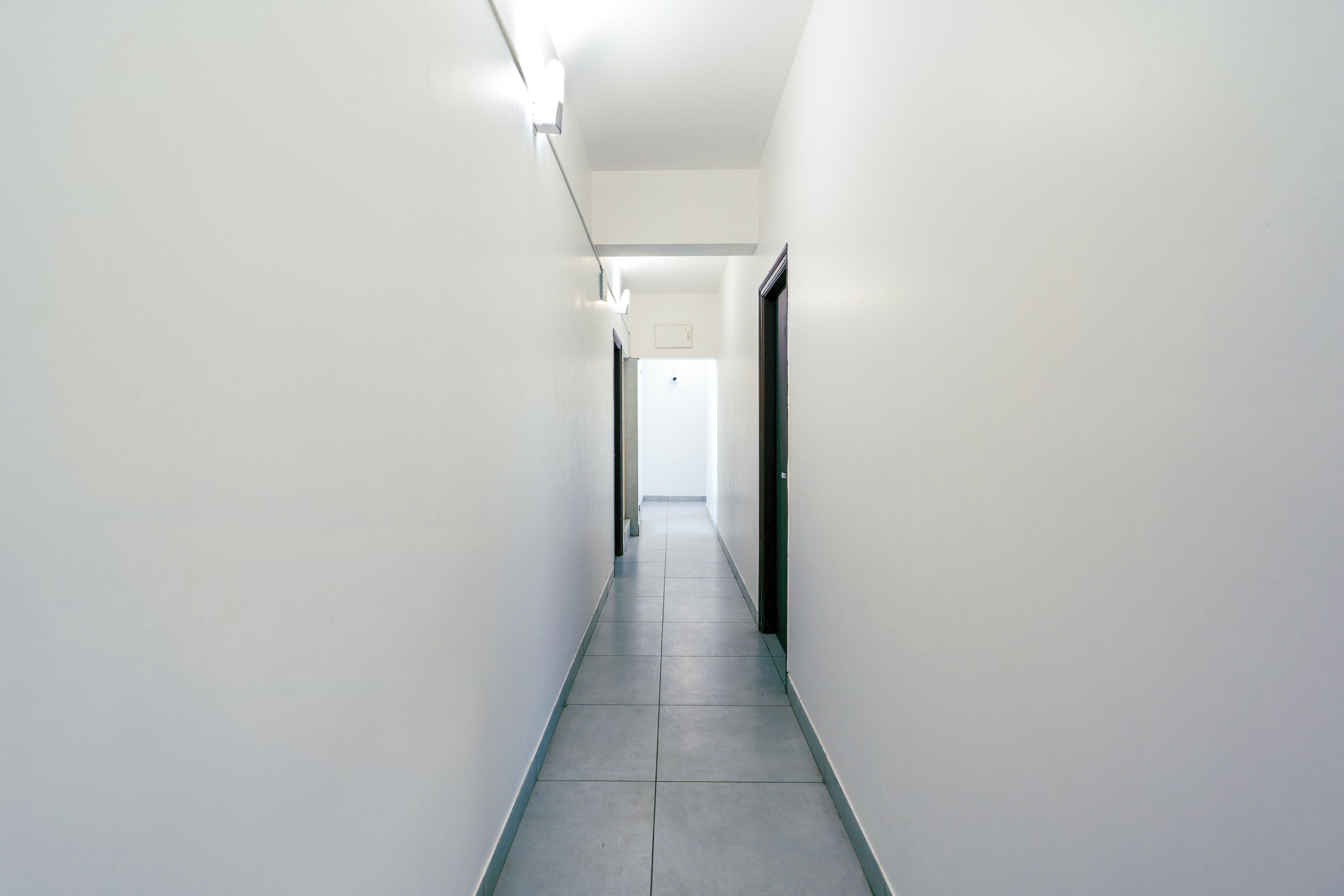 A long, narrow hallway with white walls and a tiled floor, leading to a door at the end.