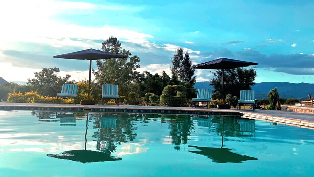 A clear swimming pool with lounge chairs and umbrellas, surrounded by trees and mountains in the background. - Molma Lodge (Greater Tubatse)