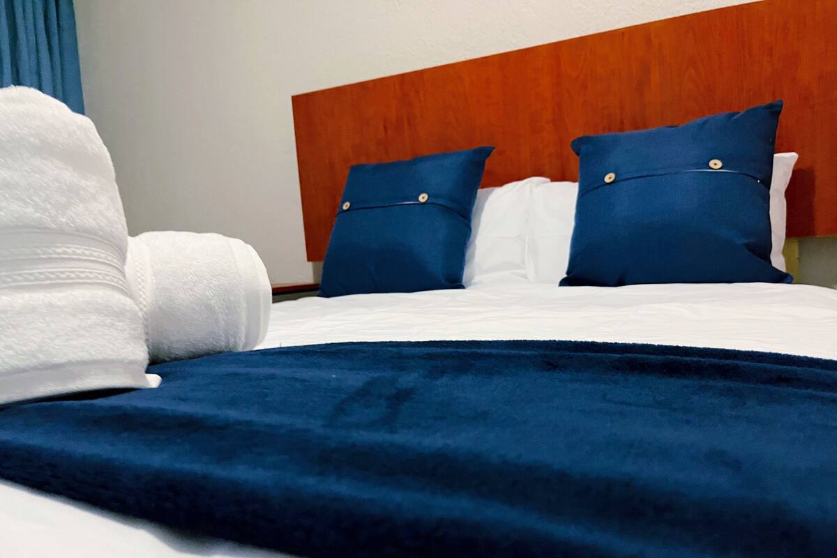 A neatly made bed with white linens, a dark blue blanket, and two matching blue pillows.