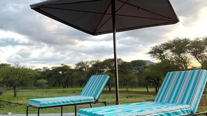 Two lounge chairs with striped cushions and a large umbrella, set on a concrete surface with a grassy field and trees in the background. - Molma Lodge (Greater Tubatse)