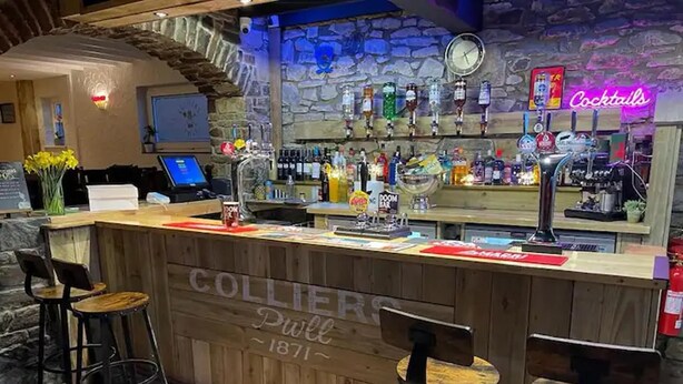 Un espace bar avec un comptoir en bois, des tabourets de bar, un panneau néon indiquant « Cocktails », ainsi que diverses bouteilles et verres.