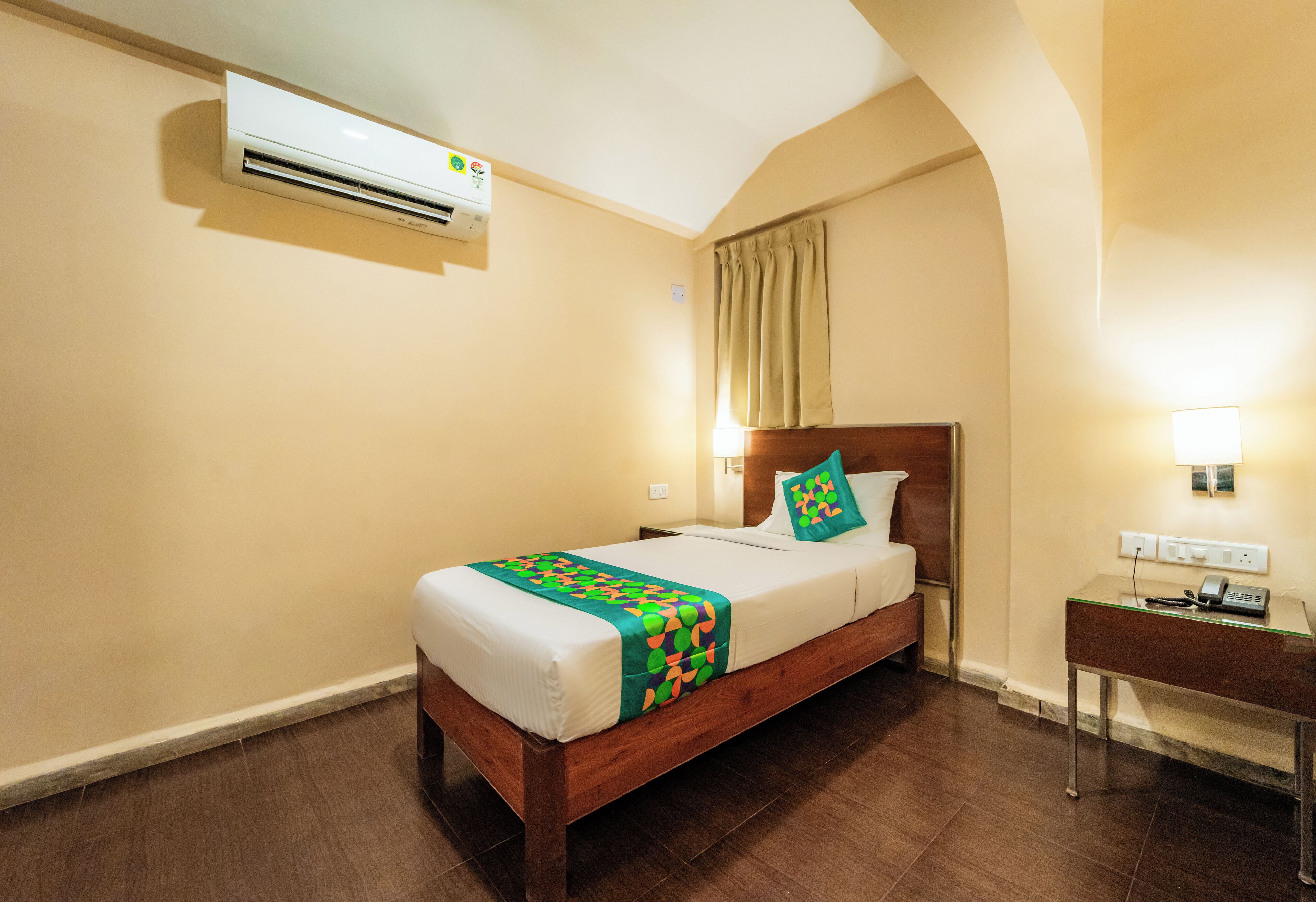 A hotel room with a wooden bed, a bedside table with a lamp, an air conditioning unit, and a small desk with a phone.