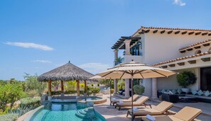 A pool area with lounge chairs and umbrellas, surrounded by greenery and a building with a tiled roof. - Stunning Ocean Views, Private Pool, & Seaside Golf (Cabo San Lucas)