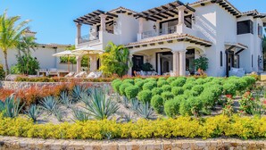 Une grande maison à étages, à l’extérieur blanc, dotée de nombreux balcons et d’un jardin bien entretenu où poussent diverses plantes et arbustes.