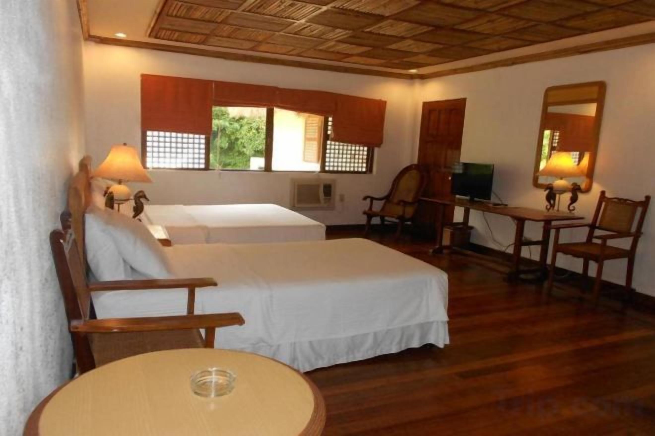 A hotel room with two beds, a round table, a chair, and a desk with a lamp.