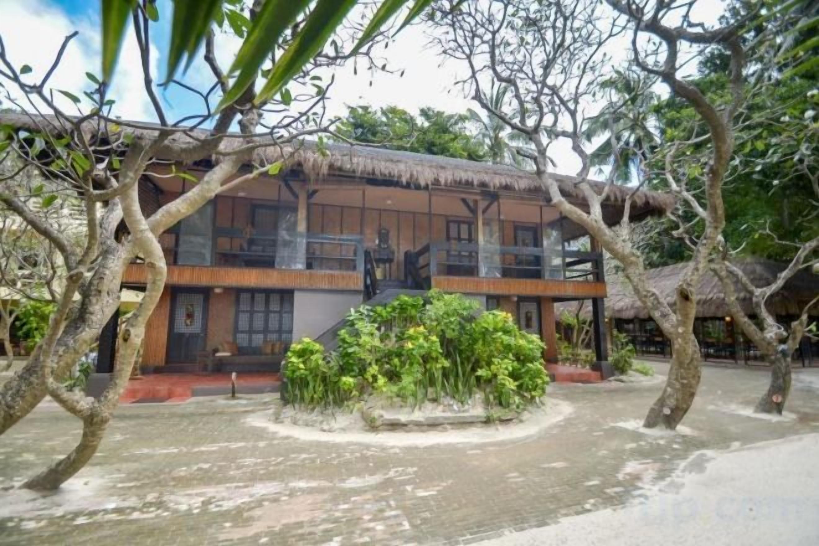 A two-story building with a thatched roof, surrounded by trees and a small garden.
