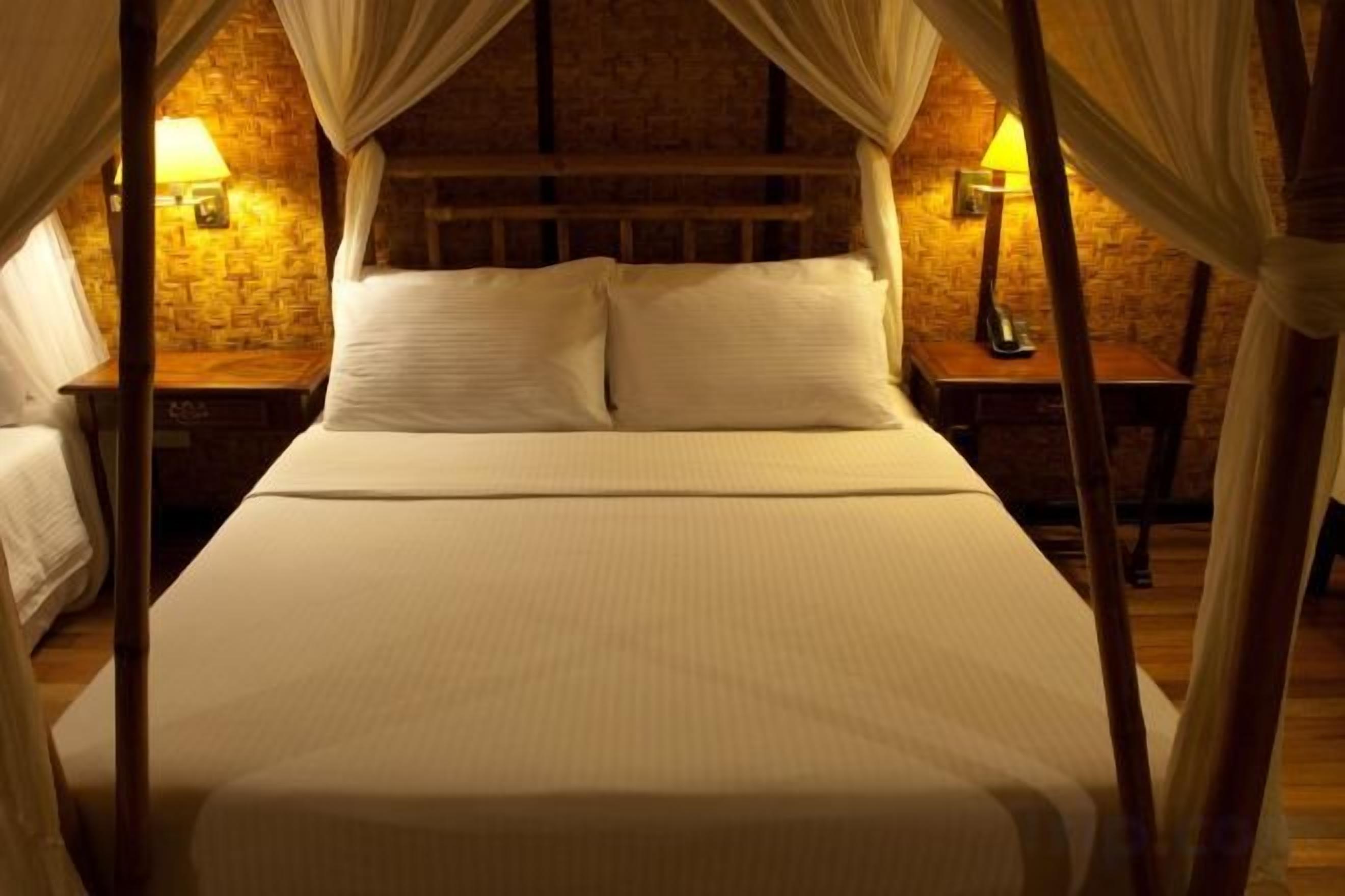 Una cama con dosel de cuatro postes, ropa de cama blanca, una pared de ladrillo y dos lámparas de noche.