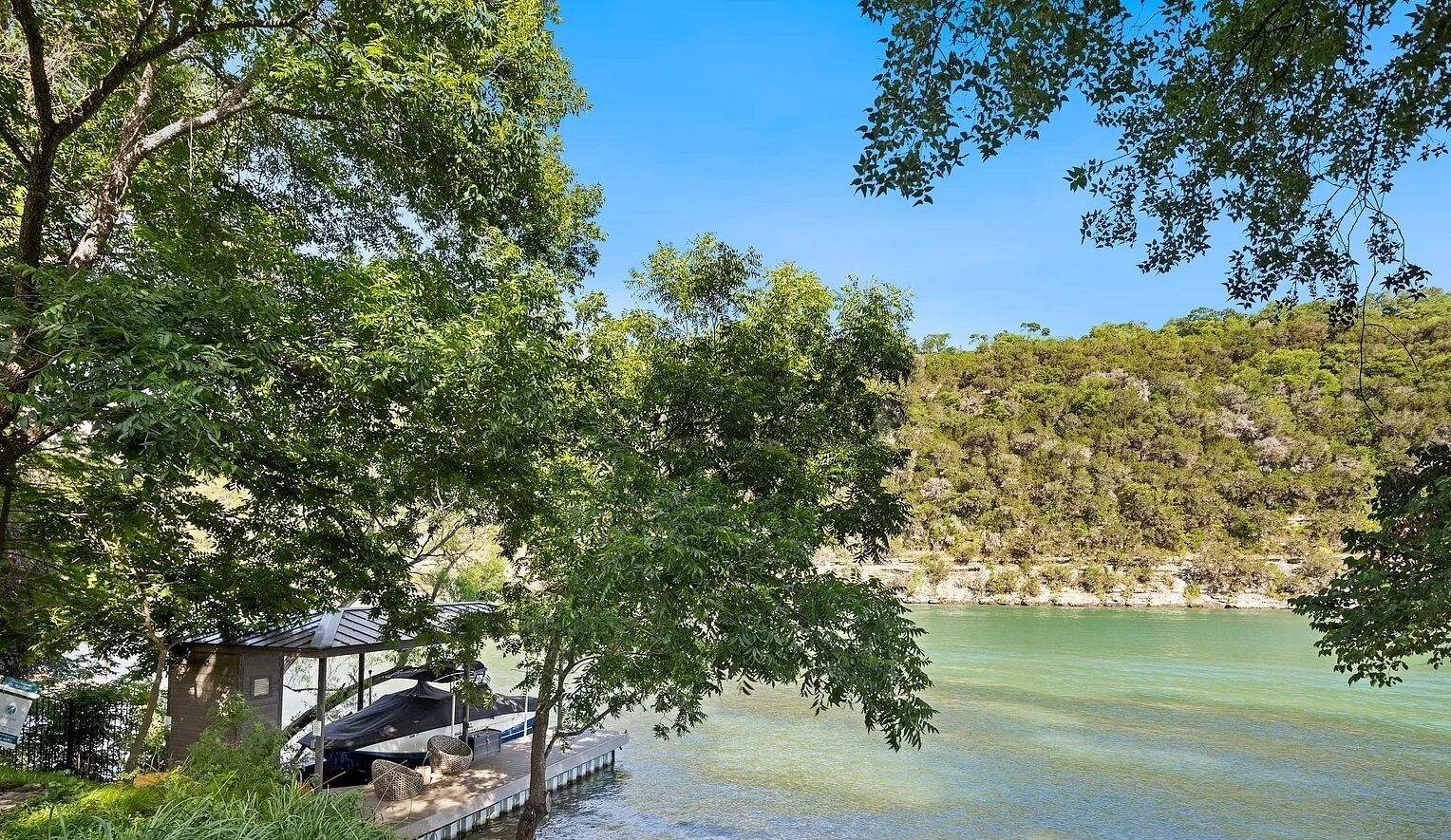 A lakeside cabin with a dock extending into the water, surrounded by lush greenery and a forested hillside.