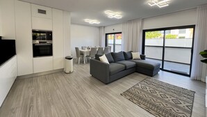 A modern living room with a grey sectional sofa, a dining table with chairs, a kitchen area with an oven, and a large window with a view of the outside. - FARO PRESTIGE by HOMING (Faro)