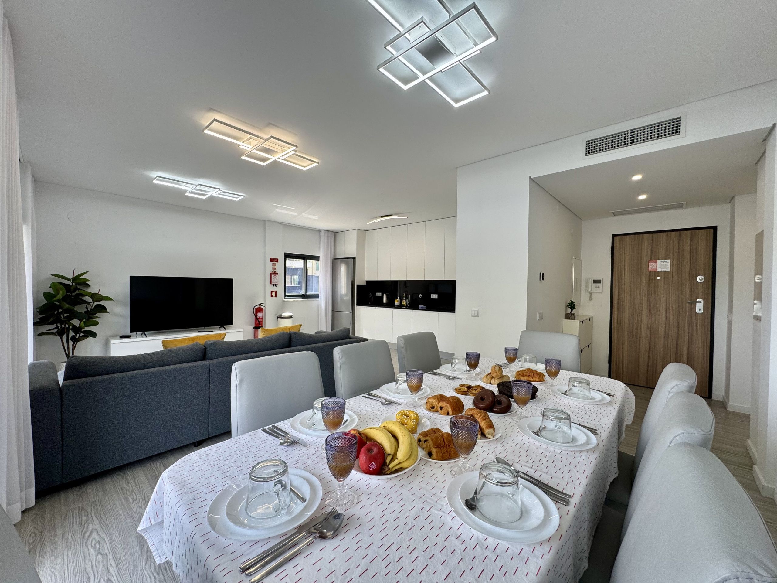 A modern dining area with a table set for breakfast, a sofa, and a TV in the background.