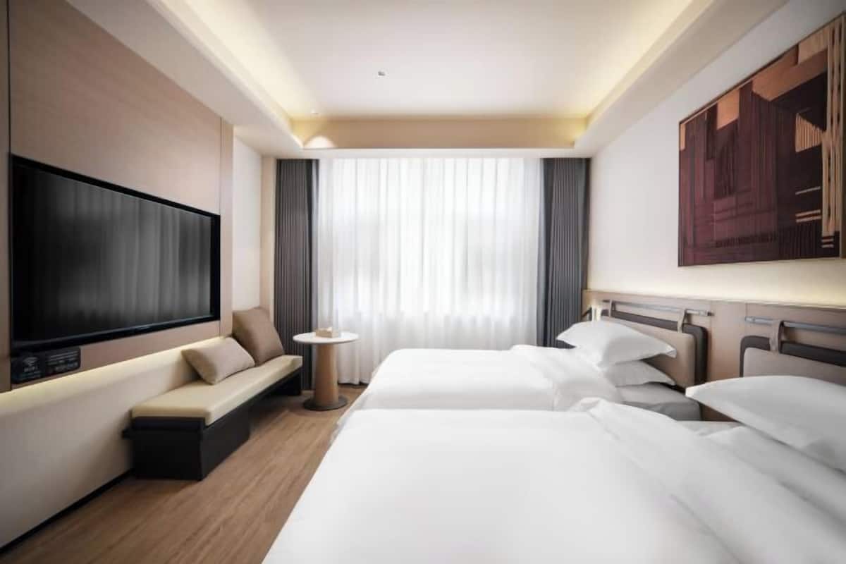 Ein Hotelzimmer mit Fernseher, zwei Betten, einer Couch, einem kleinen Tisch und einem großen Fenster mit Vorhängen.