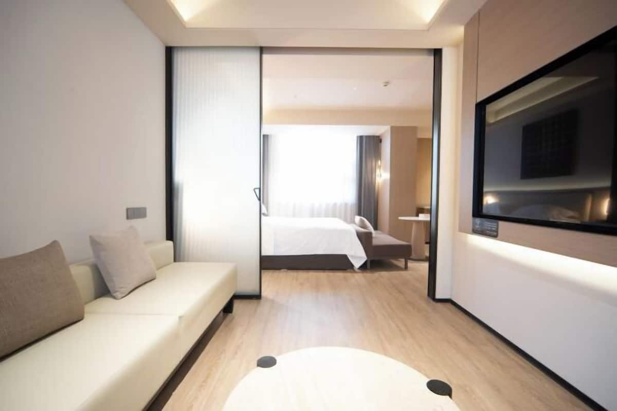 Ein modernes Hotelzimmer mit einer Couch, einem Bett, einem Fernseher und einer Schiebetür zu einem weiteren Raum.