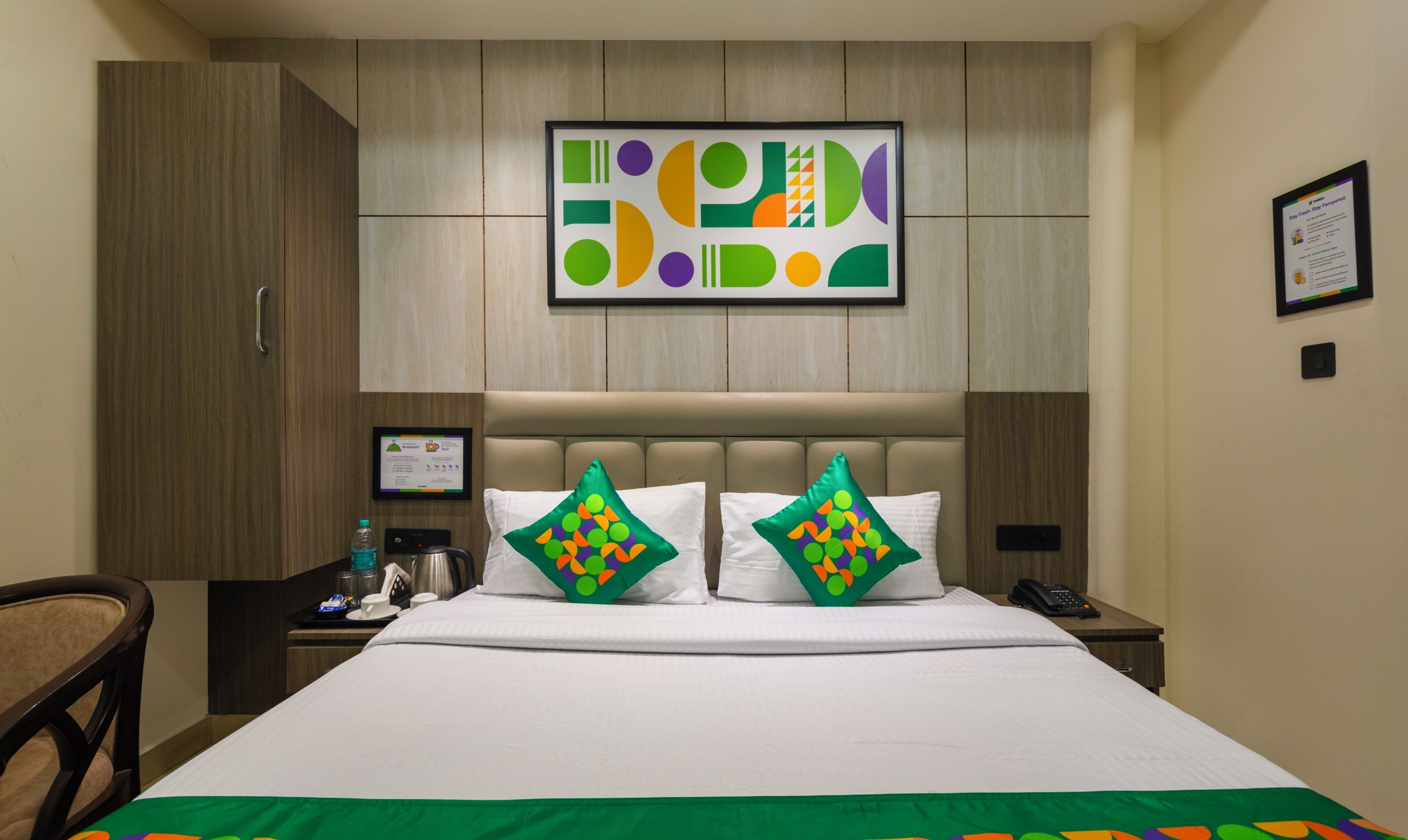 Una camera d'albergo con un letto grande, due cuscini con un motivo colorato, un comodino con un telefono e una bottiglia d'acqua, e un quadro appeso alla parete.