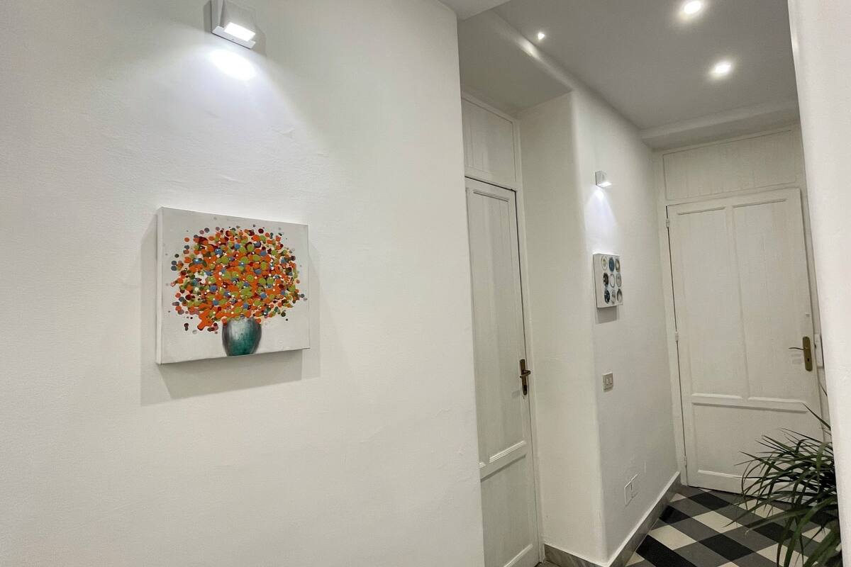 A hallway with a white door, a potted plant, and a painting of a vase with colorful flowers.