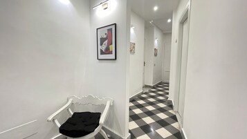 Un corridoio con pavimento a scacchi, un quadro appeso al muro e una sedia bianca con cuscino nero.