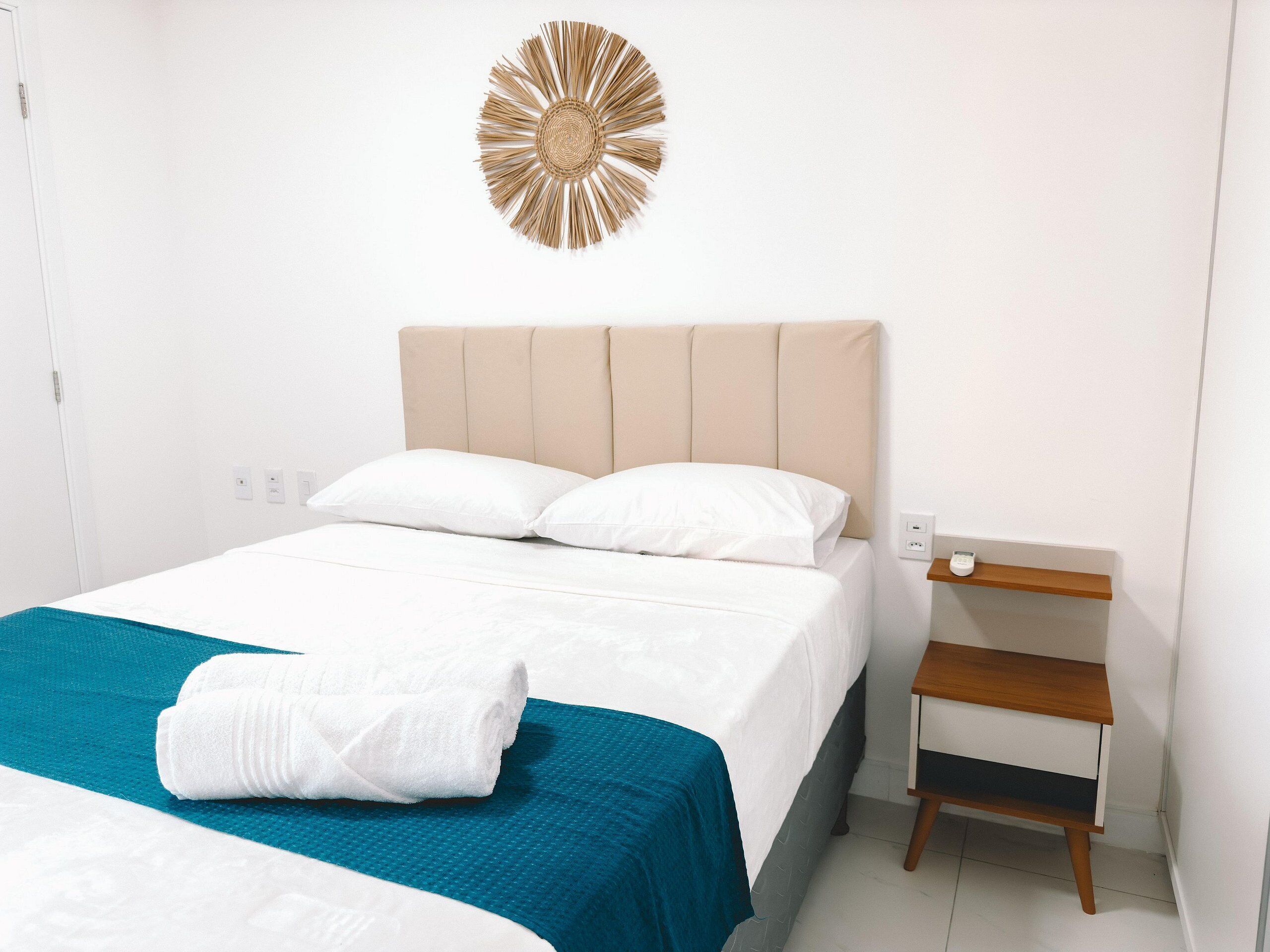 Cama bem arrumada com colcha verde-água e almofadas brancas, mesa de cabeceira de madeira com gaveta e um objeto decorativo fixado na parede.