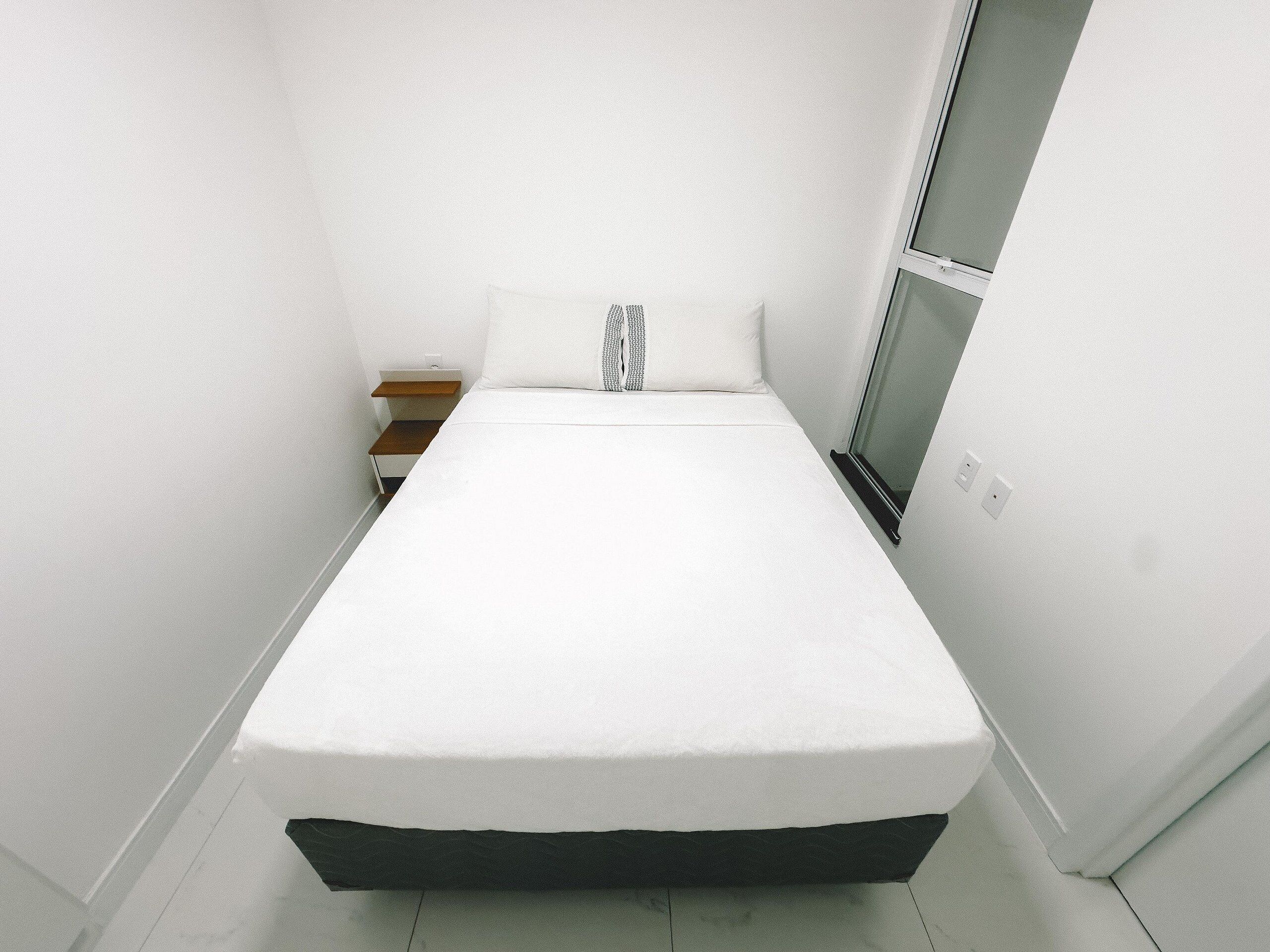 Une petite chambre blanche avec un lit simple, une table de chevet en bois, un oreiller blanc et une fenêtre avec des stores.