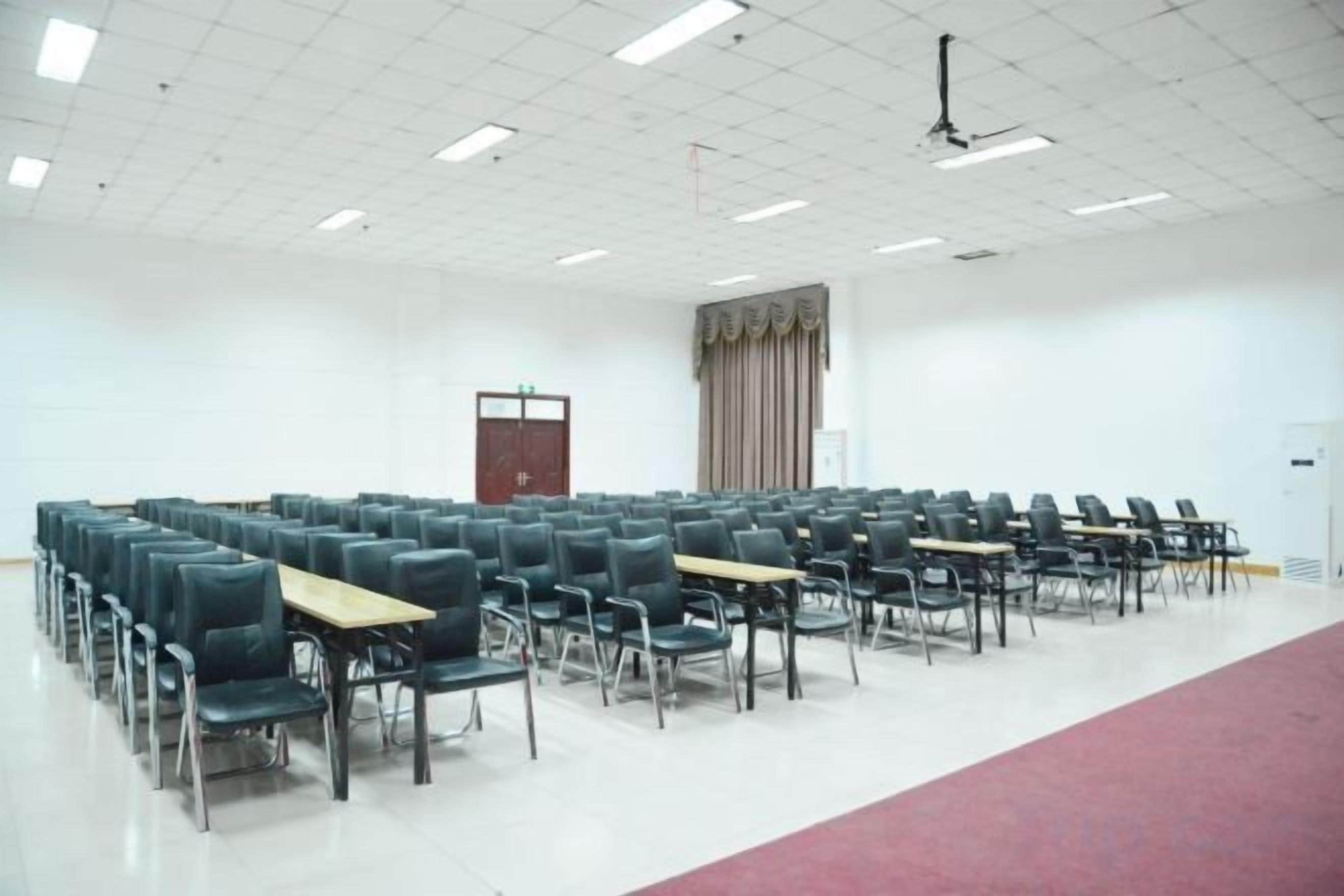 Une salle de conférence avec des rangées de chaises et de tables, un plafond blanc doté de luminaires en grille, une porte et un espace recouvert d’un tapis rouge.