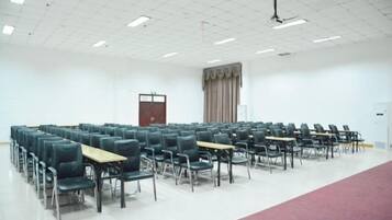 Une salle de conférence avec des rangées de chaises et de tables, un plafond blanc doté de luminaires en grille, une porte et un espace recouvert d’un tapis rouge.