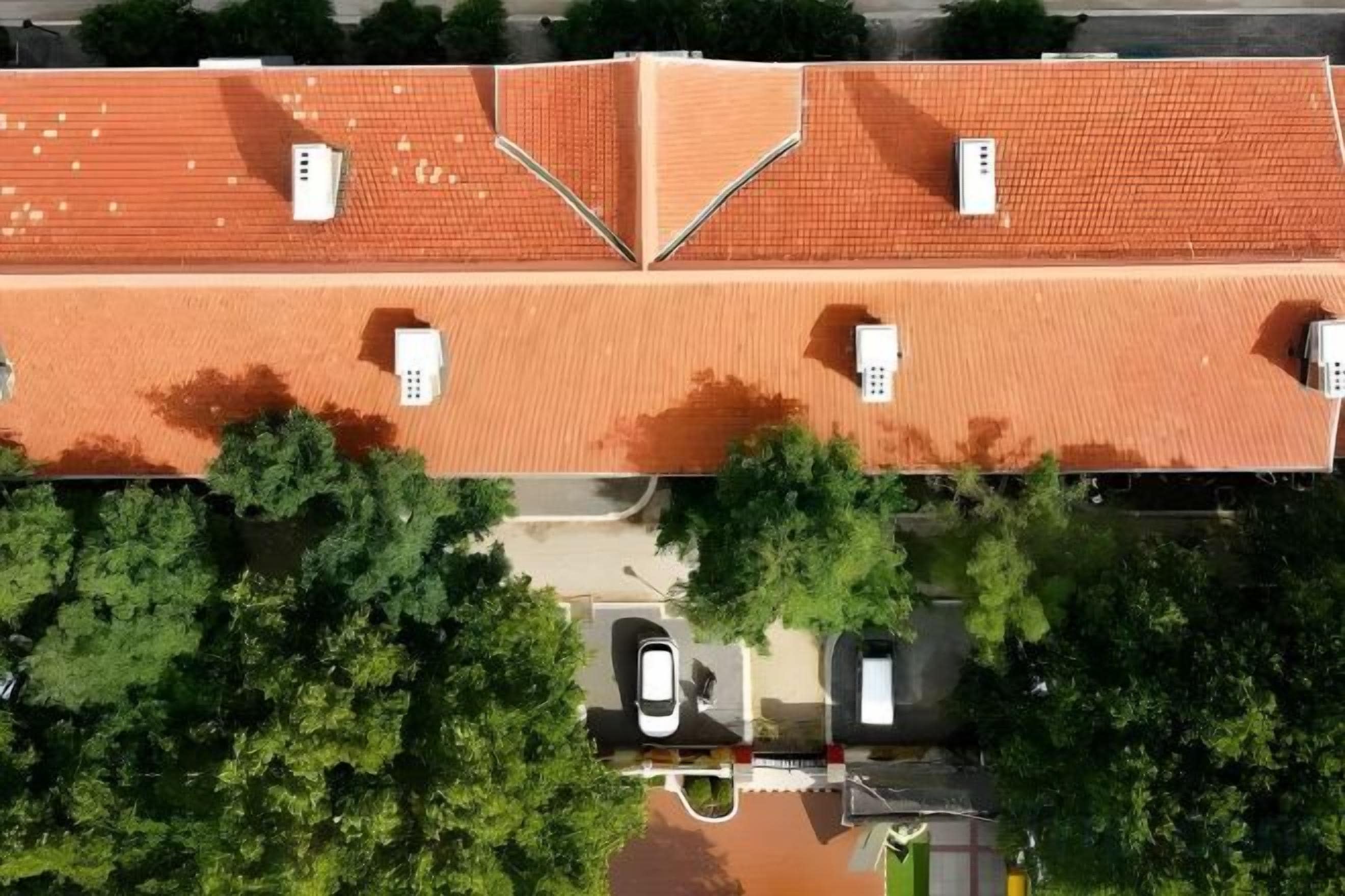 Vista aérea de un edificio con techo de tejas rojas, un área de estacionamiento y árboles en primer plano.