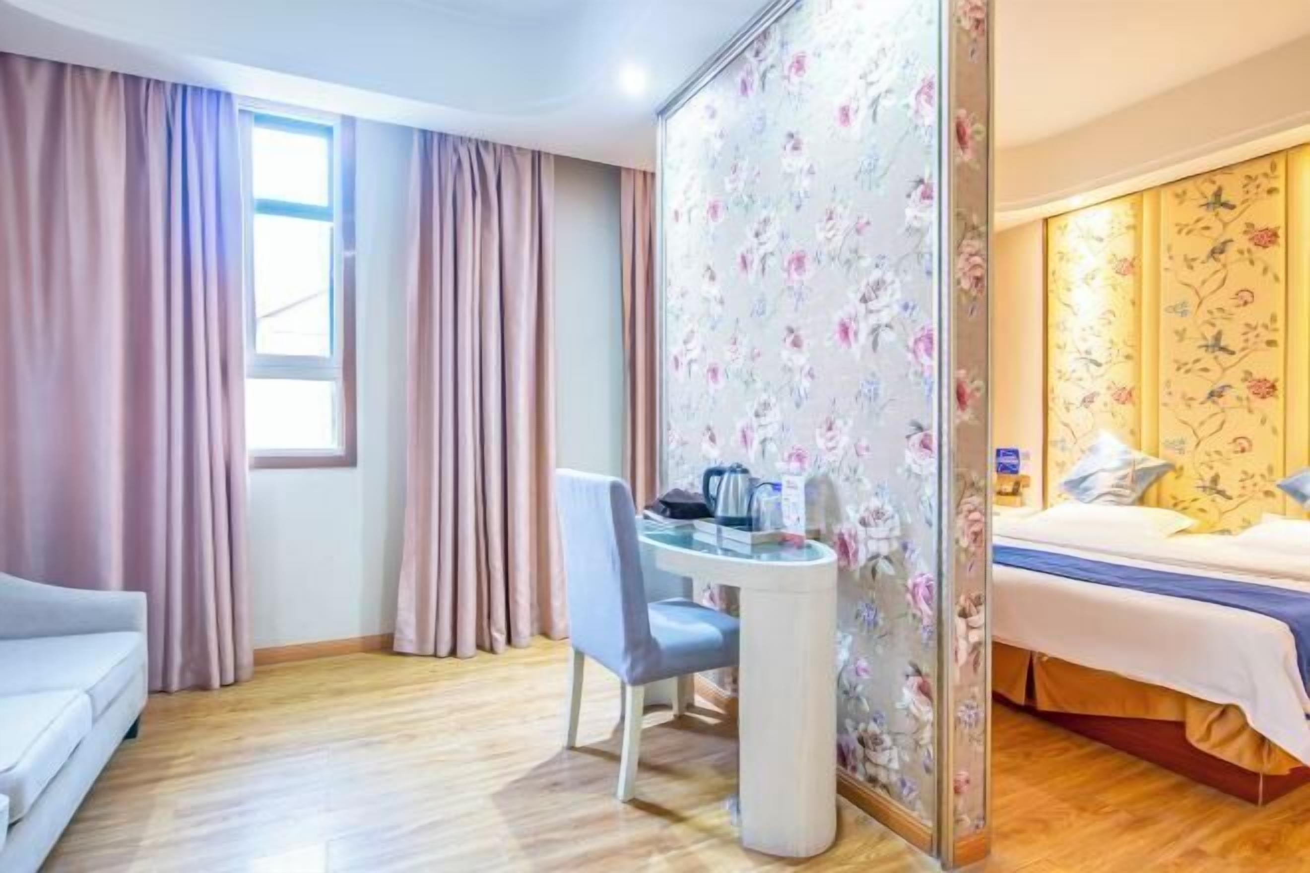 A hotel room with a bed, a desk, and a chair. There is a window with curtains, and the room has wooden flooring.