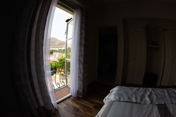 A bedroom with a bed, a balcony with a view of a mountainous landscape, and a door leading to another room.