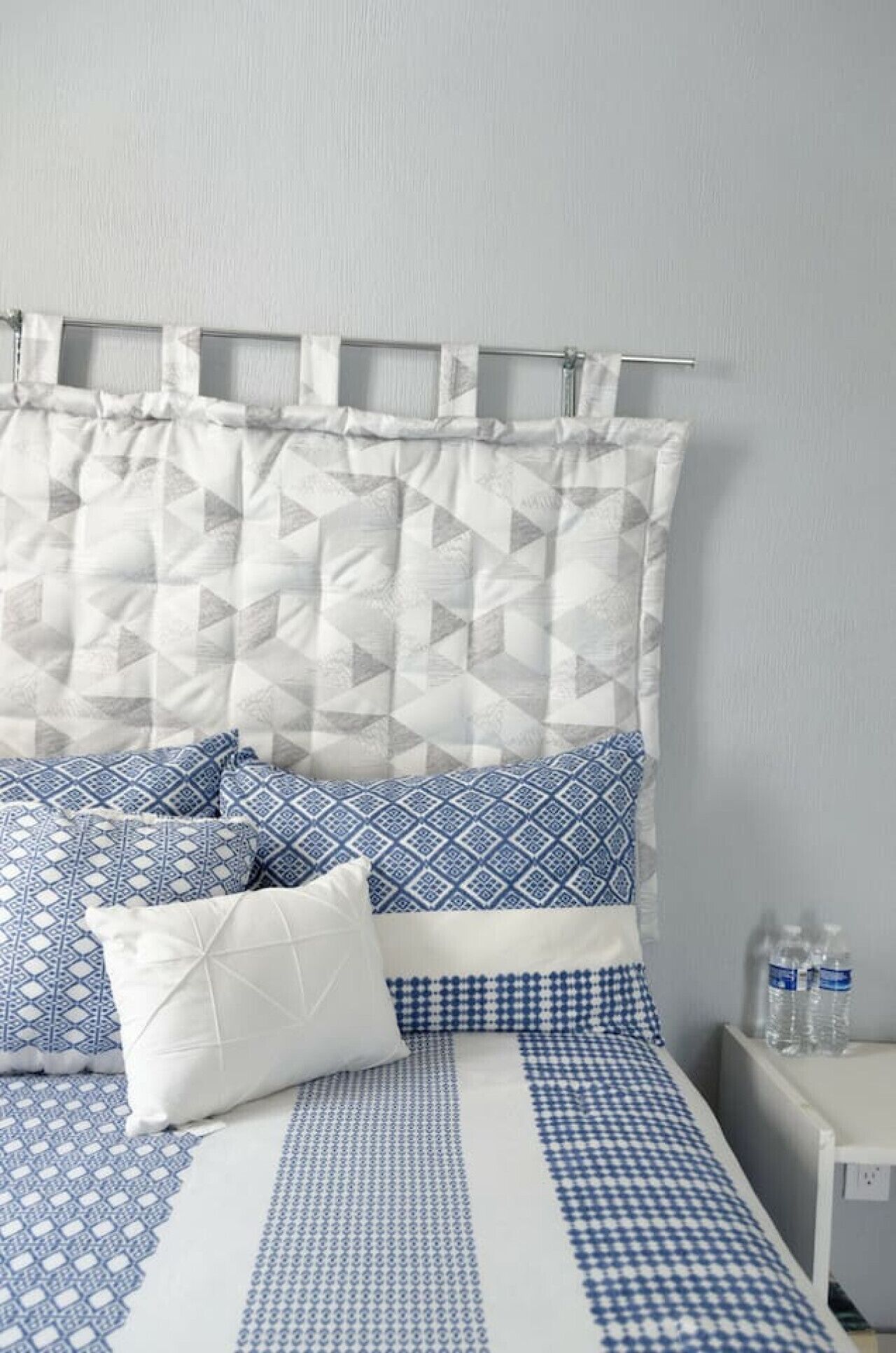 A neatly made bed with a patterned bedspread and pillows, a headboard with a geometric design, and a bedside table with water bottles.