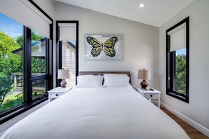 A bedroom with a large bed, two bedside tables with lamps, a window with a view of greenery, and a butterfly artwork on the wall. - Wild Goose Landing- Located near local wineries and breweries (Kingsville)