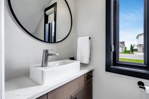 A modern bathroom with a white sink, a round mirror, and a window with a view of houses. - Wild Goose Landing- Located near local wineries and breweries (Kingsville)